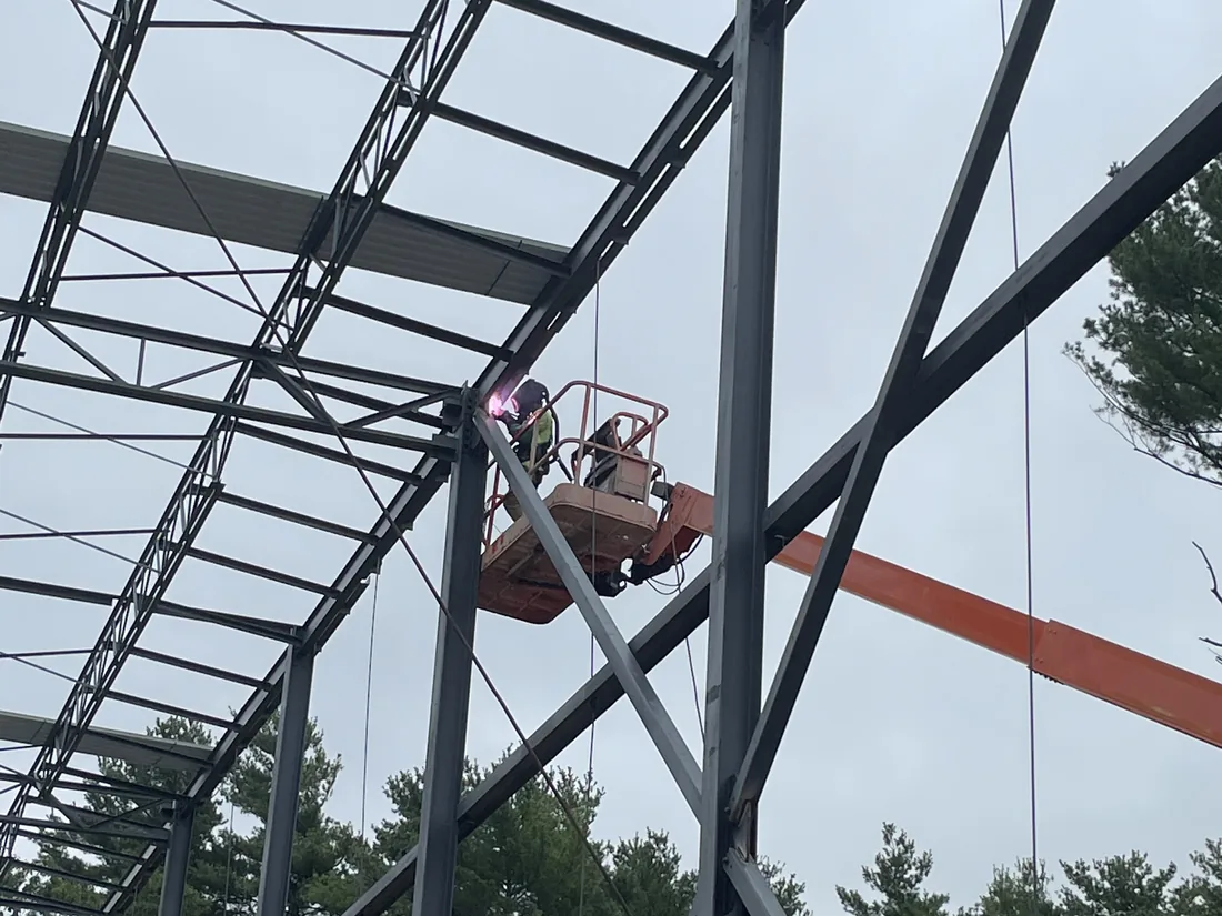 Looking up at a completed steel frame with bar joists and bridging, a boom lift working the top chord